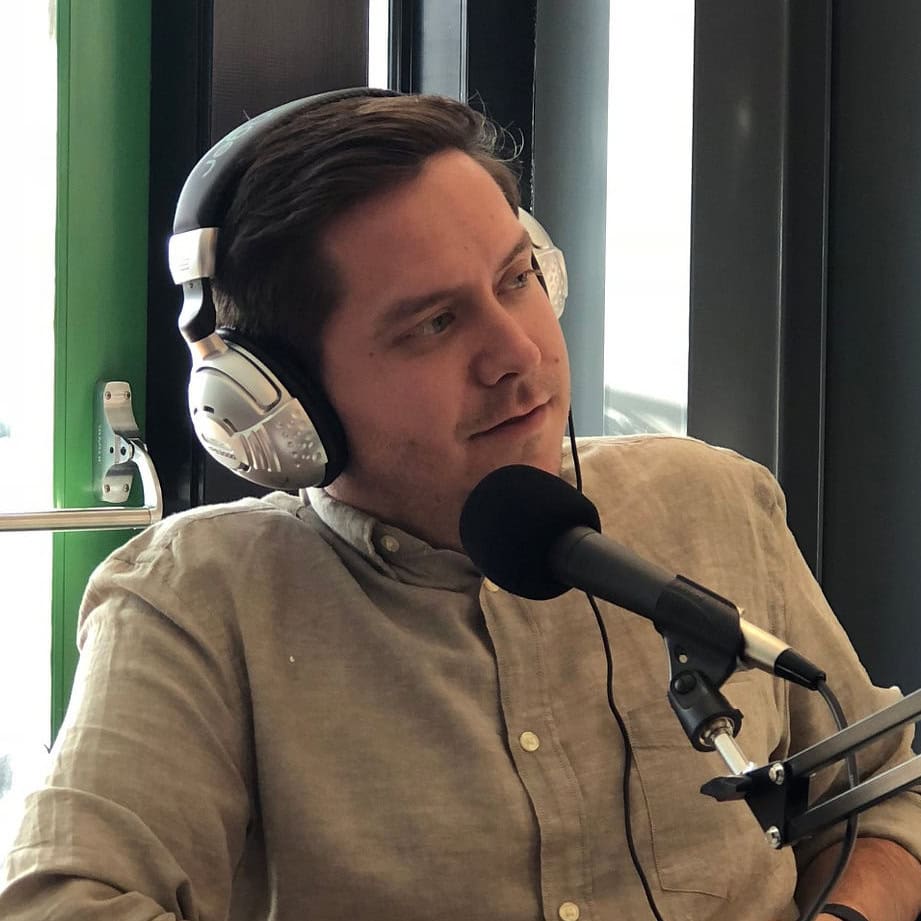 A man wearing headphones speaks into a microphone in a recording studio, sitting at a table near a window.