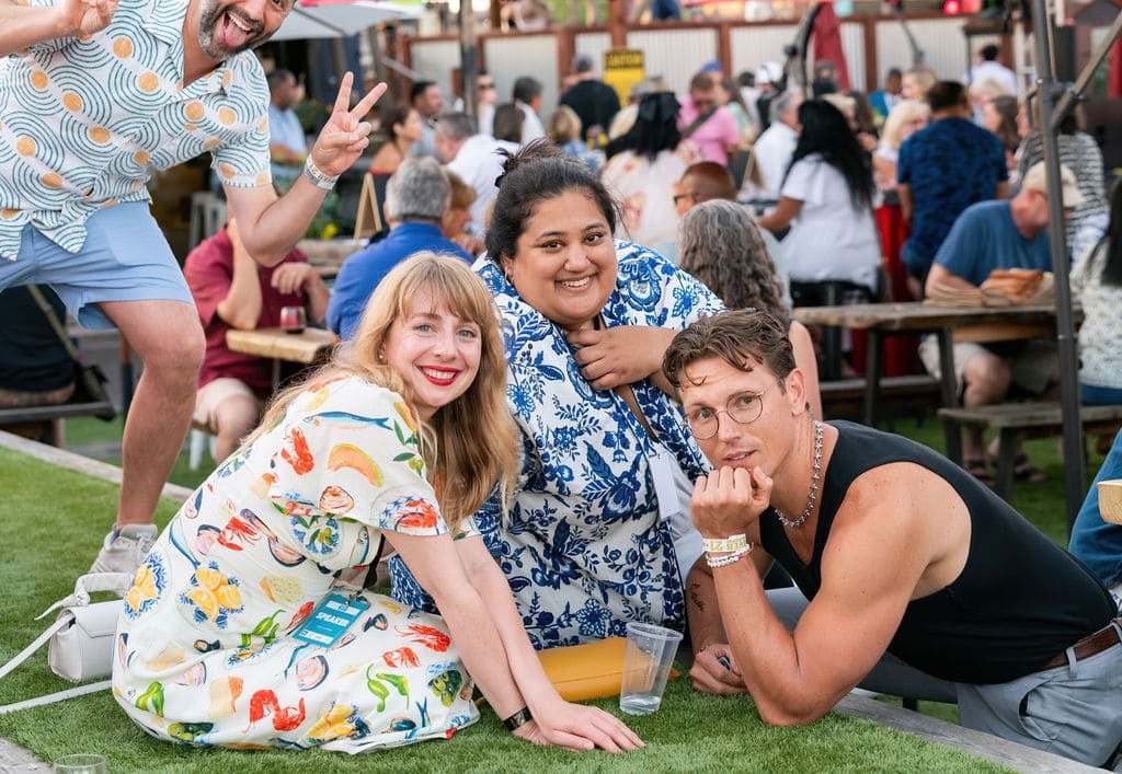 Three people posing and smiling on grass at an outdoor event, with a crowd sitting at picnic tables in the background. One person in the back is making a peace sign.
