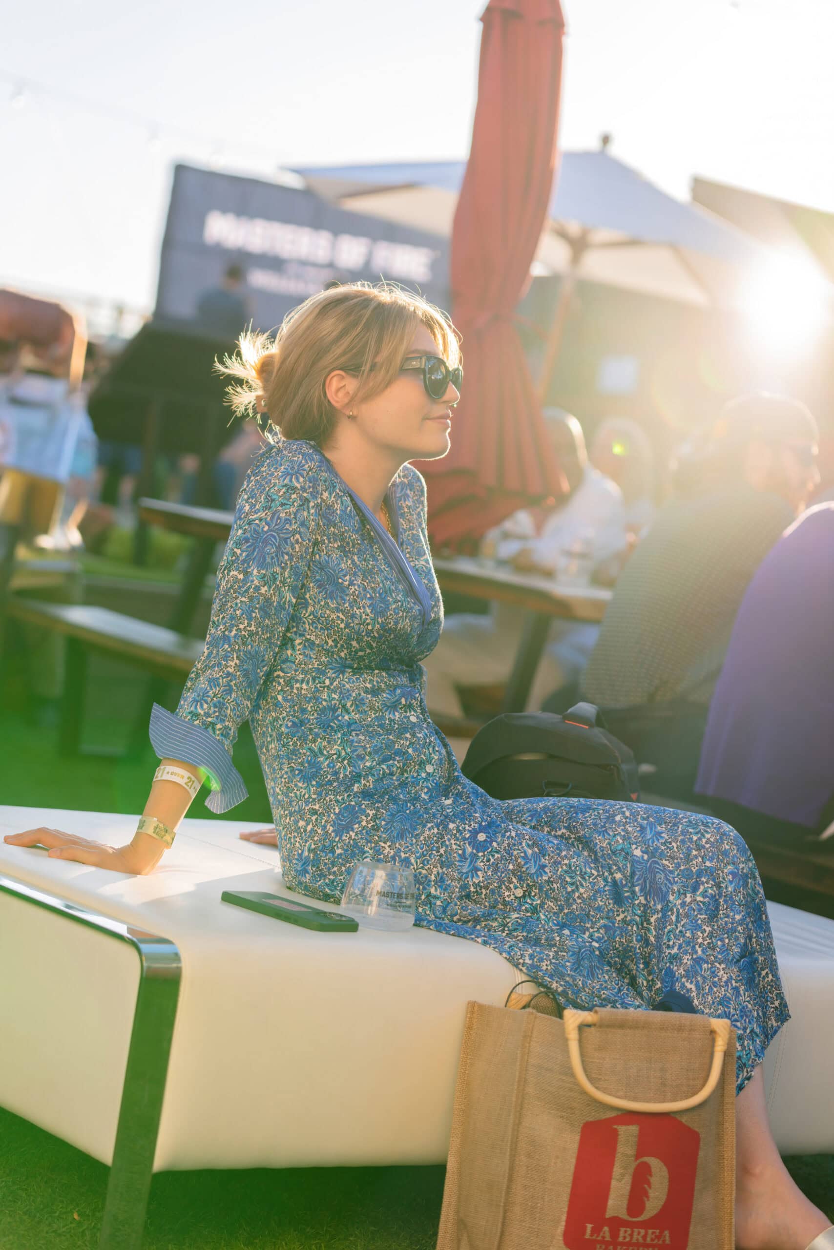 A woman in a blue floral dress and sunglasses sits on an outdoor bench, with a tote bag and a drink beside her, at a sunny outdoor gathering.