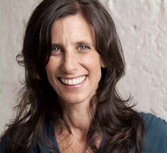A woman with long brown hair and a blue top smiles at the camera in front of a light-colored textured wall.