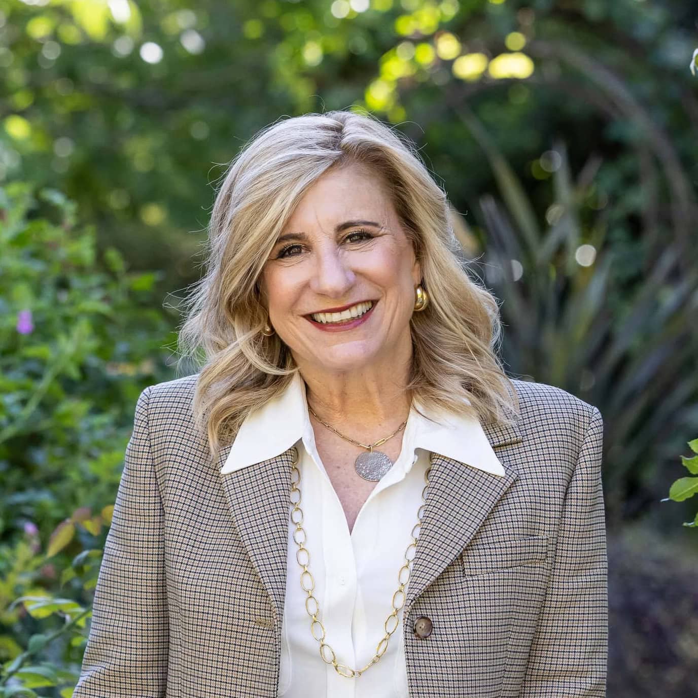 A woman with blonde hair in a checked blazer and white shirt stands outdoors, smiling, with greenery in the background.