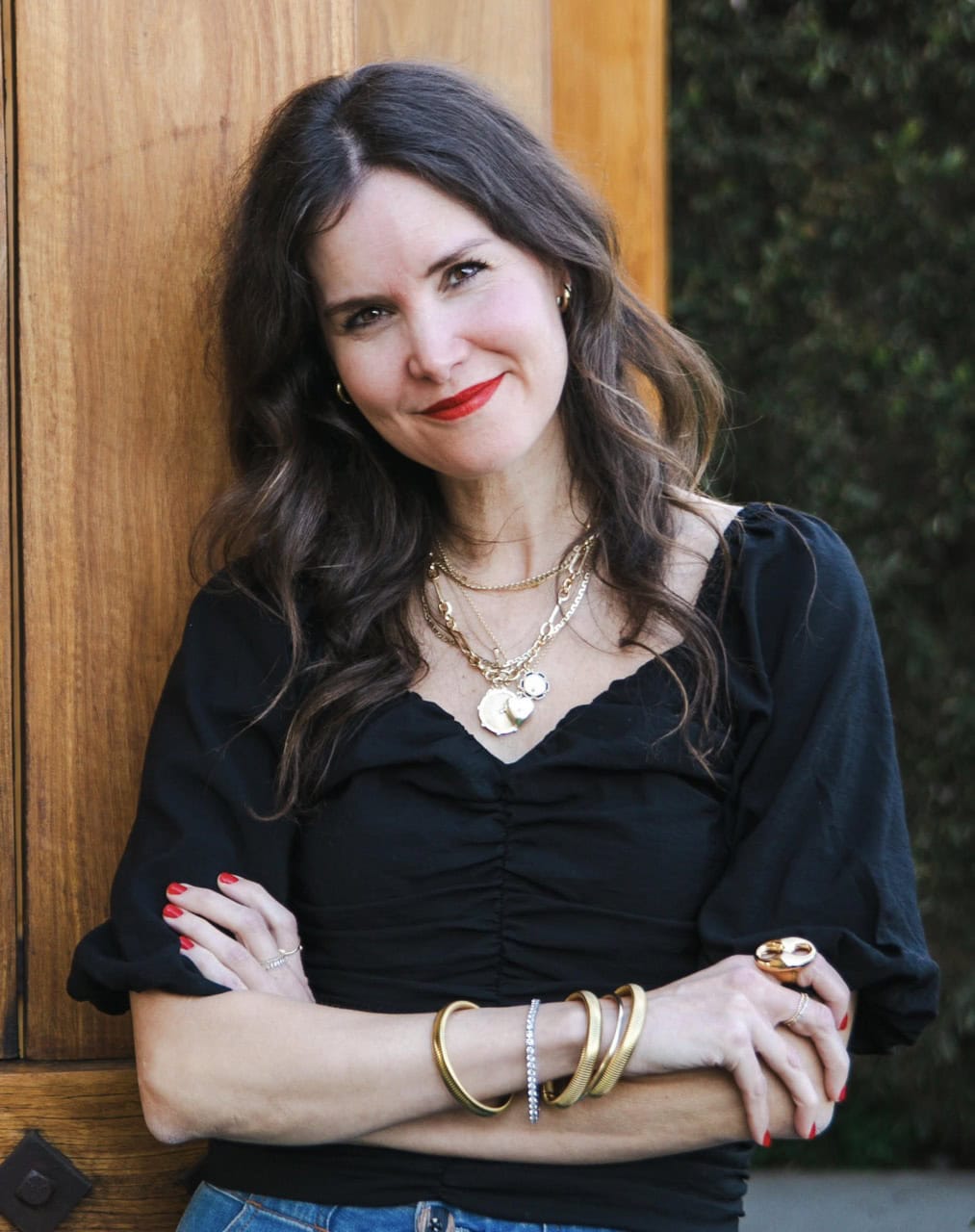 A woman with long brown hair in a black top, wearing layered gold jewelry, stands with arms crossed and smiles in front of a wooden door.